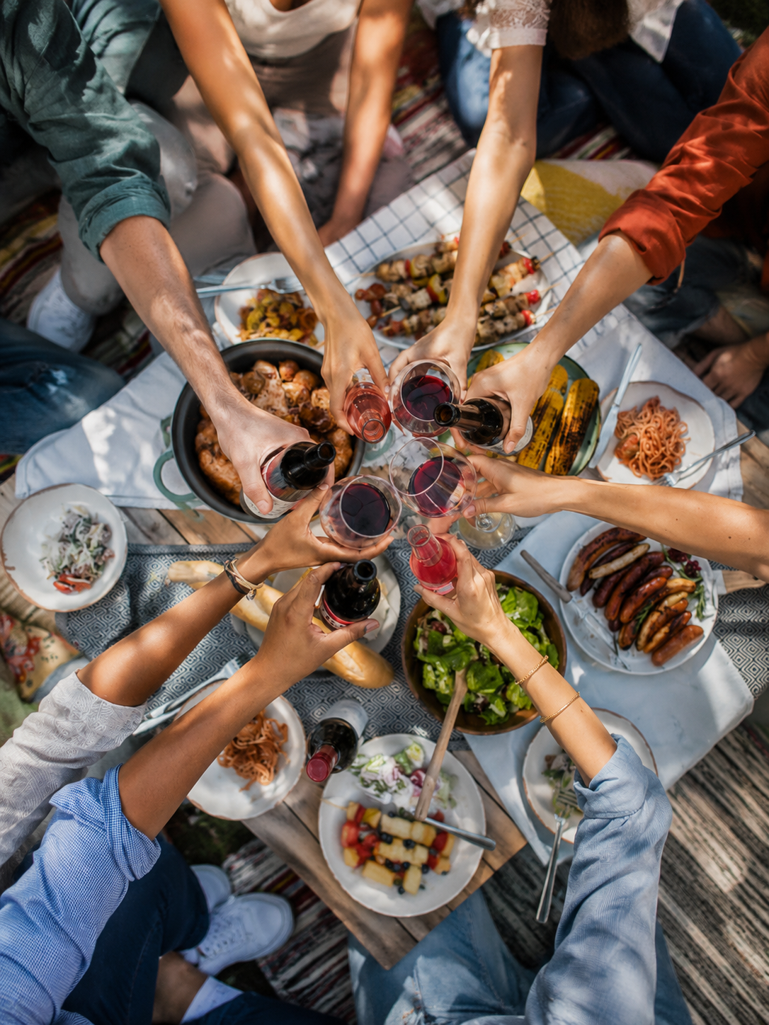  Group of friends toasting over a spread of BBQ food including meat skewers, sausages and salad at an outdoor gathering
